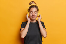 Waist Up Shot Of Pleased Young Woman Listens Music Via Headphones Keeps Eyes Closed Dressed In Casual Black T Shirt Enjoys Loud Sound Isolated Over Vivid Yellow Background. People And Hobby Concept
