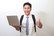 © Yazid Nasuha - Indonesian senior high school student wearing white shirt uniform with gray tie holding a laptop and showing thumb up hand gesture, standing against gray background