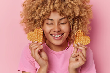 Wall Mural - Cheerful beautiful woman with curly hair holds two heart shaped waffles smiles broadly keeps eyes closed imagines how she eats this tasty food dressed casually isolated over pink background.