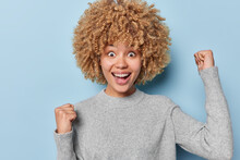Happy attractive curly haired woman clenches fists with triumph and exclaims loudly feels like winner dressed in casual grey jumper isolated over blue wall. Cheerful female model makes winner gesture
