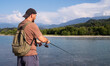 Spinning fishing. A man in a cap and with a backpack fishes with a spinning rod on a mountain river against the backdrop of mountains and a blue sky in summer on a sunny day, close-up.