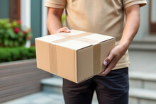 Close Up Hands Of Delivery Man Holding Parcel Box Or Cardboard Box In Front Of House Entrance. Distribution Concept Of Transportation And Delivery.