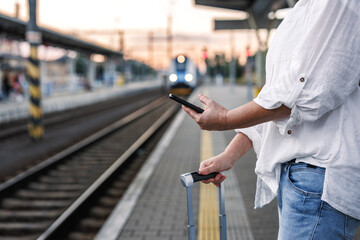 Woman with suitcase is using smartphone and mobile app to purchase online transport ticket. Tourist waiting for train at the station. Solo travel