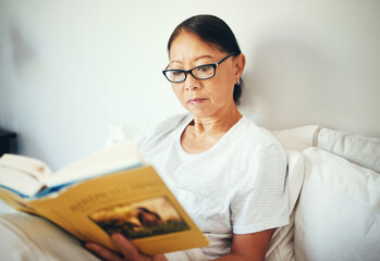 Sticker - Serious, woman and reading books in bedroom for story, fiction novel and knowledge at home. Mature asian person, glasses and comfortable in bed with literature for rest, hobby or relax to enjoy break