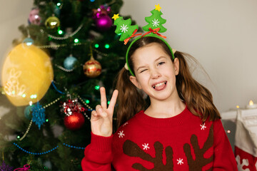 Sticker - Funny portrait of a teenage girl in a Christmas outfit against the background of a Christmas tree, looking at the camera