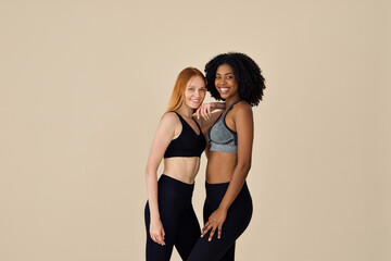 Wall Mural - Two happy diverse fit women, African and European young sporty gen z girls friends multiracial models wearing sportswear tops leggings standing smiling looking at camera on beige background.