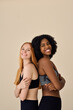 Two happy diverse fit women, African and European young sporty gen z girls friends models wearing sportswear tops leggings standing back to back smiling looking at camera on beige background.