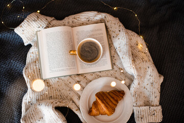 Wall Mural - Open paper book with folded pages in heart shape and cup of coffee with croissant on knitted sweater over Christmas lights in room close up. Winter cozy home atmosphere.