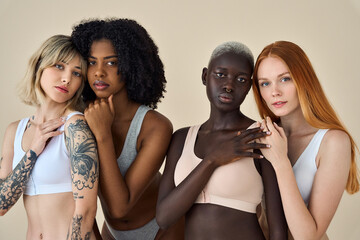 Wall Mural - Confident diverse women, four multicultural young girls friends international beauty models group wearing underwear bonding looking at camera standing isolated on beige background, portrait.