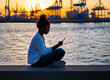 Eine junge Frau mit dunklen hochgebundenen Haaren sitzt auf einer Mauer und schaut auf ihr Smartphone, seitlicher Anblick, im Hintergrund Hafenkräne im Sonnenuntergang, horizontal, KI-generiert