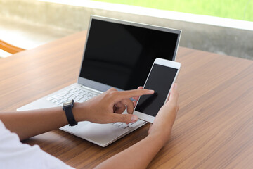 Close up of businessman hand touching blank screen of mobile phone with laptop beside on the table.  Display of smartphone and laptop mock up