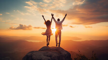 Silhouette Of Couple Jumping On Top Of A Rock And Raising Hands Up To The Sky At Beautiful Sunset.