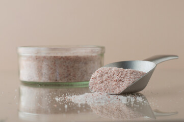 Himalayan pink salt in a glass container with a metal spatula on a beige background on a reflective surface, horizontal.