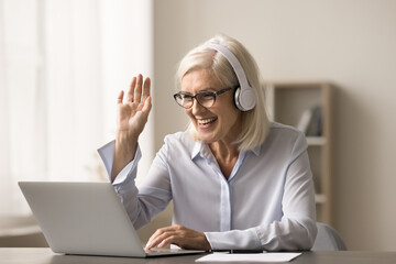 Wall Mural - Happy blonde senior student attending online educational conference. Cheerful teacher giving webinar. Positive business woman meeting with colleagues on Internet, waving hand hello on video call