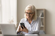 © fizkes - Happy blonde retired senior business woman typing on mobile phone at home work table, using software, online application, Internet service for job communication, sitting at laptop, smiling