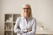 © fizkes - Cheerful confident blonde elderly business woman in elegant glasses posing indoors, standing with folded arms, looking at camera, laughing. Female professional, leader front portrait