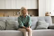 Concerned blonde senior woman sitting on couch in modern home interior, looking at window away, thinking on problems, bad anxious news, suffering from stress, feeling nervous