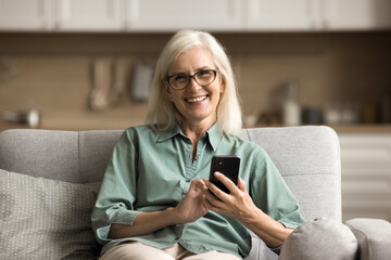 Canvas Print - Happy blonde senior woman in stylish glasses using application, online service on mobile phone for getting medical consultation on Internet, looking at camera with toothy smile, sitting on sofa