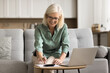© fizkes - Positive blonde elderly woman in eyeglasses calculating investment income, savings at laptop, counting insurance benefit, enjoying work on personal finance, domestic budget