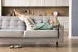 Cheerful sleepy blonde older lady lying on couch, leaning legs and feet on soft back, enjoying leisure, lazy break in modern home interior, breathing fresh cool pure air with closed eyes