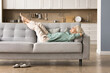© fizkes - Cheerful sleepy blonde older lady lying on couch, leaning legs and feet on soft back, enjoying leisure, lazy break in modern home interior, breathing fresh cool pure air with closed eyes