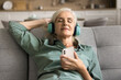 © fizkes - Peaceful relaxed senior lady enjoying music, comfort on soft couch at home, using mobile phones and big headphones, listening to online ambient retro playlist for chill, relaxation