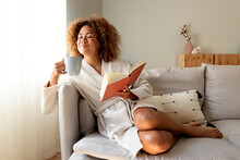 Pensive relaxed Multiracial hispanic woman holding a book, drinking morning coffee sitting on the sofa.