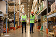 © alfa27 - Caucasian engineers in safety helmets and green uniforms walking among shelves with goods in warehouse talking, checking goods. logistic and business export, distribution and inventory concept