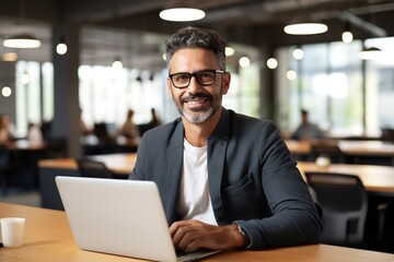 Portrait of middle-age smiling handsome business man using laptop computer, typing, working in modern office looking at camera