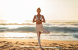 © Prostock-studio - Cheerful young european lady in sportswear practicing yoga on sea beach, enjoying workout outdoor on nature, full length