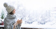 © scharfsinn86 - A woman in a warm hat and blanket holds smartphone in her hands. Winter landscape with snowfall in the background
