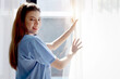 © Stella - Side view of happy smiling beautiful woman doctor with stethoscope in blue coat standing and opening white curtain at window in clinic hospital. Young female nurse and medical health care worker.