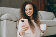 © ViDi Studio - Young smiling happy woman in casual clothes sits near grey sofa couch use mobile cell phone drink coffee stay at home hotel flat rest relax spend free spare time in living room indoor Lounge concept