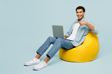 Wall Mural - Full body young IT man of African American ethnicity in shirt casual clothes sit in bag chair hold use work point on laptop pc computer isolated on plain pastel light blue background studio portrait