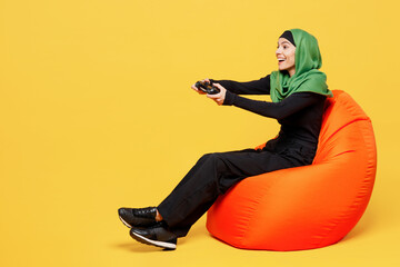 Poster - Full body young asian muslim woman wear green hijab abaya black clothes sit in bag chair hold play pc game with joystick console isolated on plain yellow background People uae islam religious concept