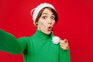 Wall Mural - Close up young woman wear green turtleneck Santa hat posing do selfie shot pov mobile cell phone point finger camera you isolated on plain red background Happy New Year 2024 Christmas holiday concept