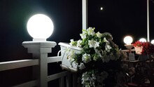 Lantern Shining On Dark Night And White Blooming Flowers Petunia In White Pot Swaying In Wind Hanging On Fence Near Table In Cafe. Romantic Atmosphere At Night In Cafe. Decor Interior Details Outdoors