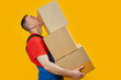 Middle-aged man in workers uniform holds large heavy boxes. Portrait of loader in studio on yellow background. Side view. Copy space, mock up.