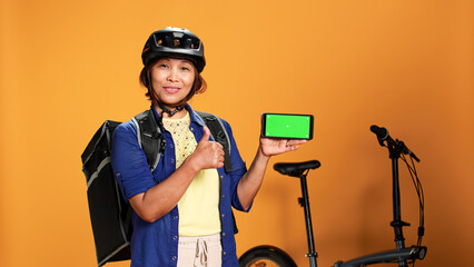 Courier holding mock up green screen smartphone, showing thumbs up hand gesturing. Delivery woman presenting chroma key touchscreen of phone in landscape mode, isolated over orange studio background