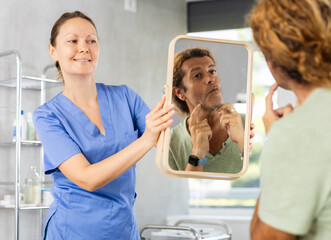 Focused man carefully examining his face after plastic surgery in mirror held by smiling female surgeon..