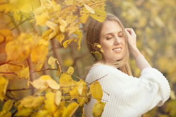 Wall Mural - Beautiful young woman with light brown hair in a white sweater on a background of foliage in an autumn park. Portrait of an attractive happy woman among colorful yellow leaves. Fall season. Autumn.