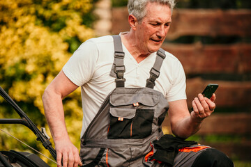 Senior man in a business suit encountering lawnmower problems in his garden, using his phone to reach out for assistance