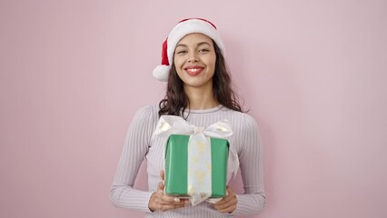 Sticker - Young beautiful hispanic woman smiling confident wearing christmas hat holding gift over isolated pink background