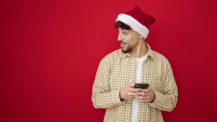 Poster - Young arab man wearing christmas hat using smartphone over isolated red background