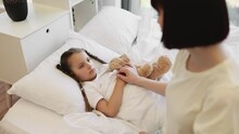 Upset caucasian girl lying in bed with her toy teddy bear while caring mother sitting near and checking temperature. Dark haired woman holding hand on child forehead during domestic treatment.