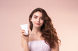 © SOLDATOOFF - Beauty portrait of an attractive brown haired woman with long hair holding a tube of cream on pastel pink background.