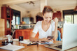 © Geber86 - Happy young woman looking at a smartphone while going over paperwork in the kitchen