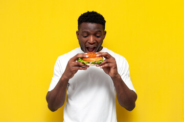 Wall Mural - african american man in white t-shirt holding big burger and biting it, the guy eats fast food with his mouth open