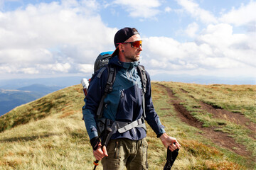 young man in orange jacket and glasses walks in the mountains with trekking stick, tourist guy with 