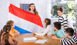 © JackF - Preteens pupils looking at national flag of Netherlands while learning different cultures with positive young female teacher in schoolroom